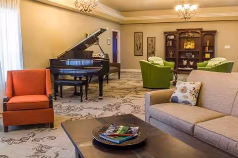 A spacious lounge with a grand piano, colorful seating, and a bookcase in a senior facility.
