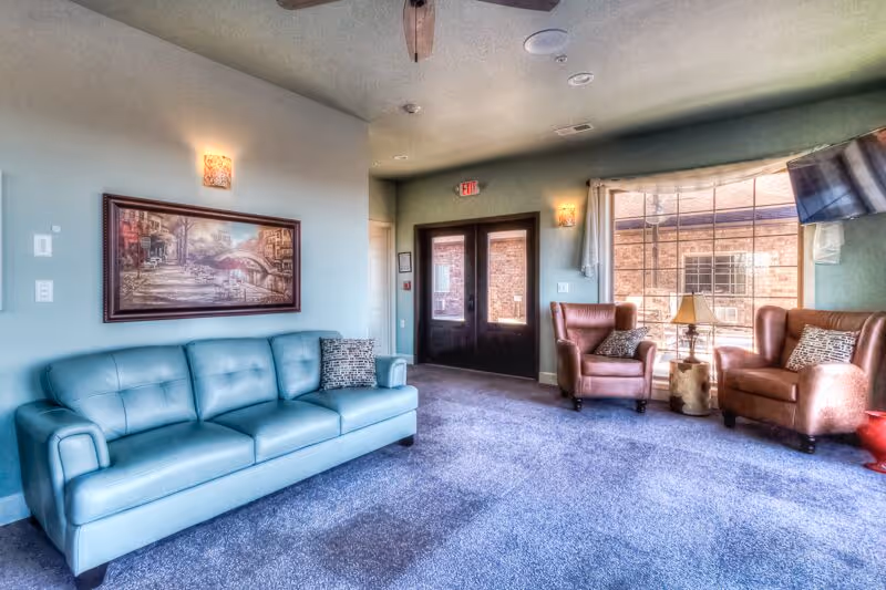 A senior living facility common area with a light blue sofa on the left, two brown leather armchairs on the right near a large window, a small wooden side table with a lamp between the chairs, a wall painting above the sofa, and double glass doors in the background.