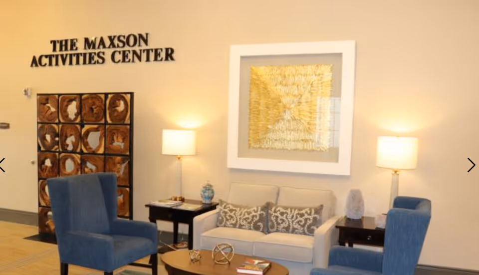 A furnished activities room with a sofa, blue armchairs, side tables and lamps beneath a wall sign reading 'The Maxson Activities Center'.