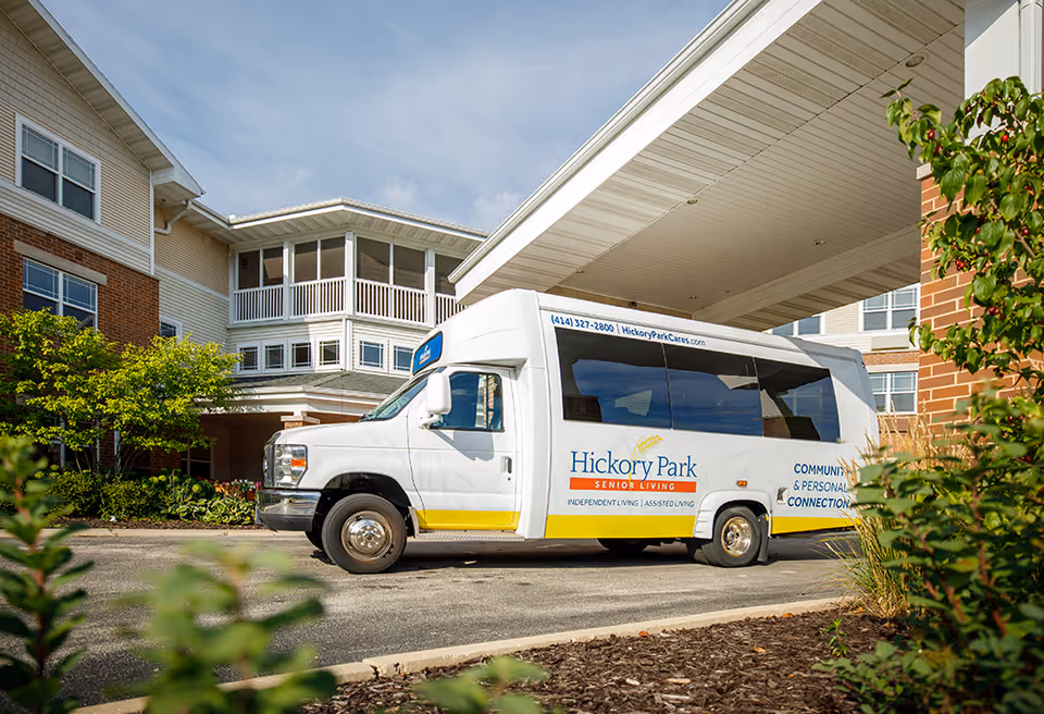 A white shuttle van with Hickory Park Senior Living branding is parked under a covered entrance of a senior living facility building with brick and beige siding, surrounded by greenery and trees.