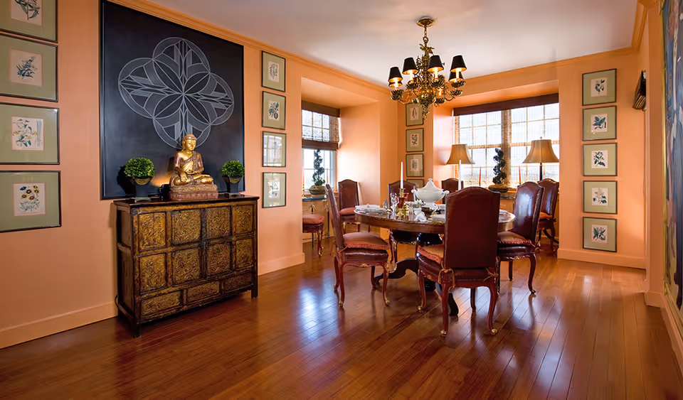Elegant dining room with a round wooden table and upholstered chairs under a chandelier, a decorative sideboard topped with a Buddha statue, and framed artwork on peach-colored walls.
