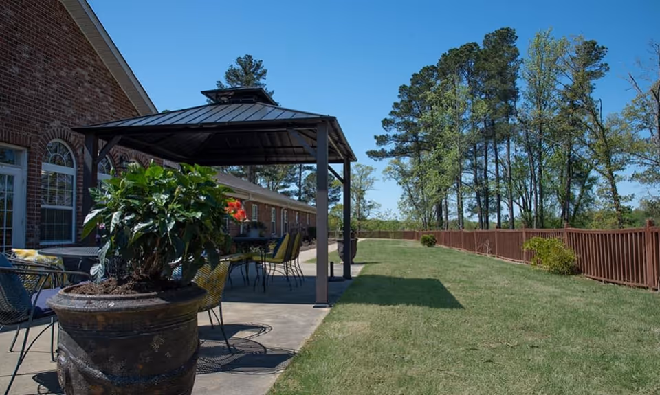 Outdoor patio area with a metal gazebo, several chairs and tables, a large potted plant in the foreground, a brick building on the left, and a grassy lawn bordered by a wooden fence and tall trees under a clear blue sky.