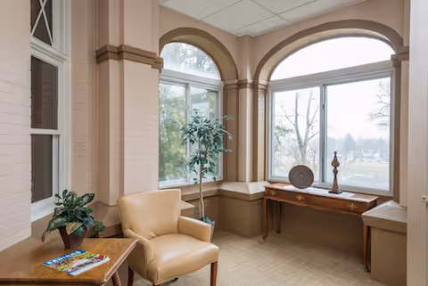 A cozy sitting area with a beige armchair, a small wooden table with magazines and a plant, a tall potted plant, and a wooden console table with decorative items placed in front of large arched windows overlooking trees and an outdoor view.