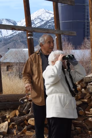 An elderly couple outdoors near a rustic wooden structure and stacked firewood. The woman is using binoculars to look into the distance while the man stands beside her holding a walking cane. Snow-capped mountains and a silo are visible in the background.