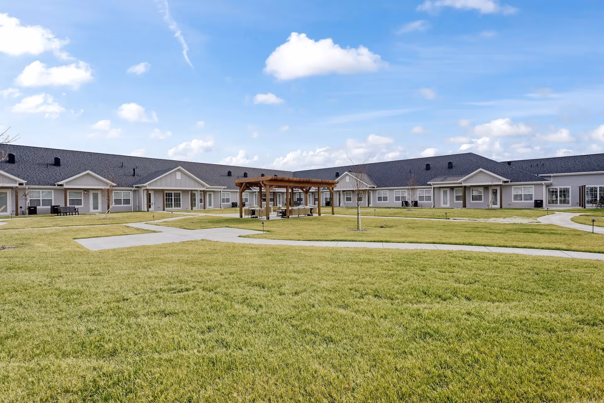 Outdoor view of Cedarhurst Senior Living of Salina showing a large grassy courtyard with paved walkways and a wooden pergola with seating in the center. The single-story building with multiple doors and windows surrounds the courtyard under a blue sky with scattered clouds.