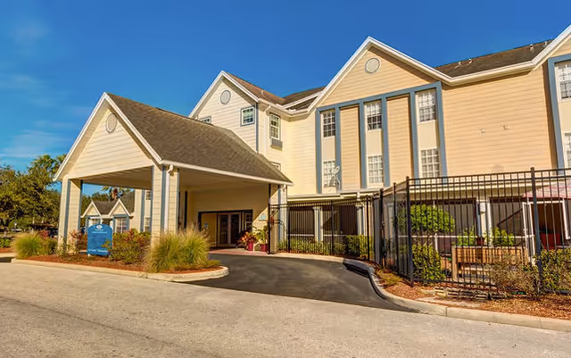 Front entrance and covered porte-cochere of a beige multi-story senior living building with a fenced patio and landscaping under a blue sky.
