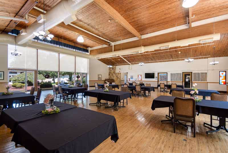 A spacious dining room with multiple tables covered in black tablecloths and chairs arranged around them. The room has large windows with white blinds, wooden floors, and a wooden ceiling with exposed beams and hanging light fixtures. There are small flower arrangements on the tables and a stone fireplace in the background.