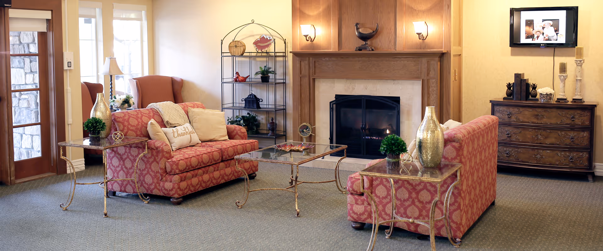A cozy senior living facility common area with two patterned red sofas facing each other, a glass coffee table in between, and a fireplace with a wooden mantel. There is a metal shelving unit with decorative items, a wooden dresser with candles and books, a wall-mounted TV, and a door with a stone wall visible outside. The room is warmly lit with wall sconces and a table lamp.
