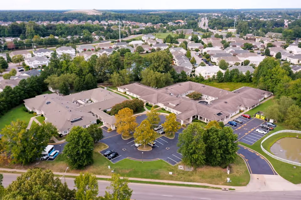 Aerial view of Chesapeake Place Senior Living facility surrounded by trees and residential neighborhoods. The building has a large parking lot with several cars parked, and a small pond is visible on the right side. The area is lush with greenery and well-maintained landscaping.