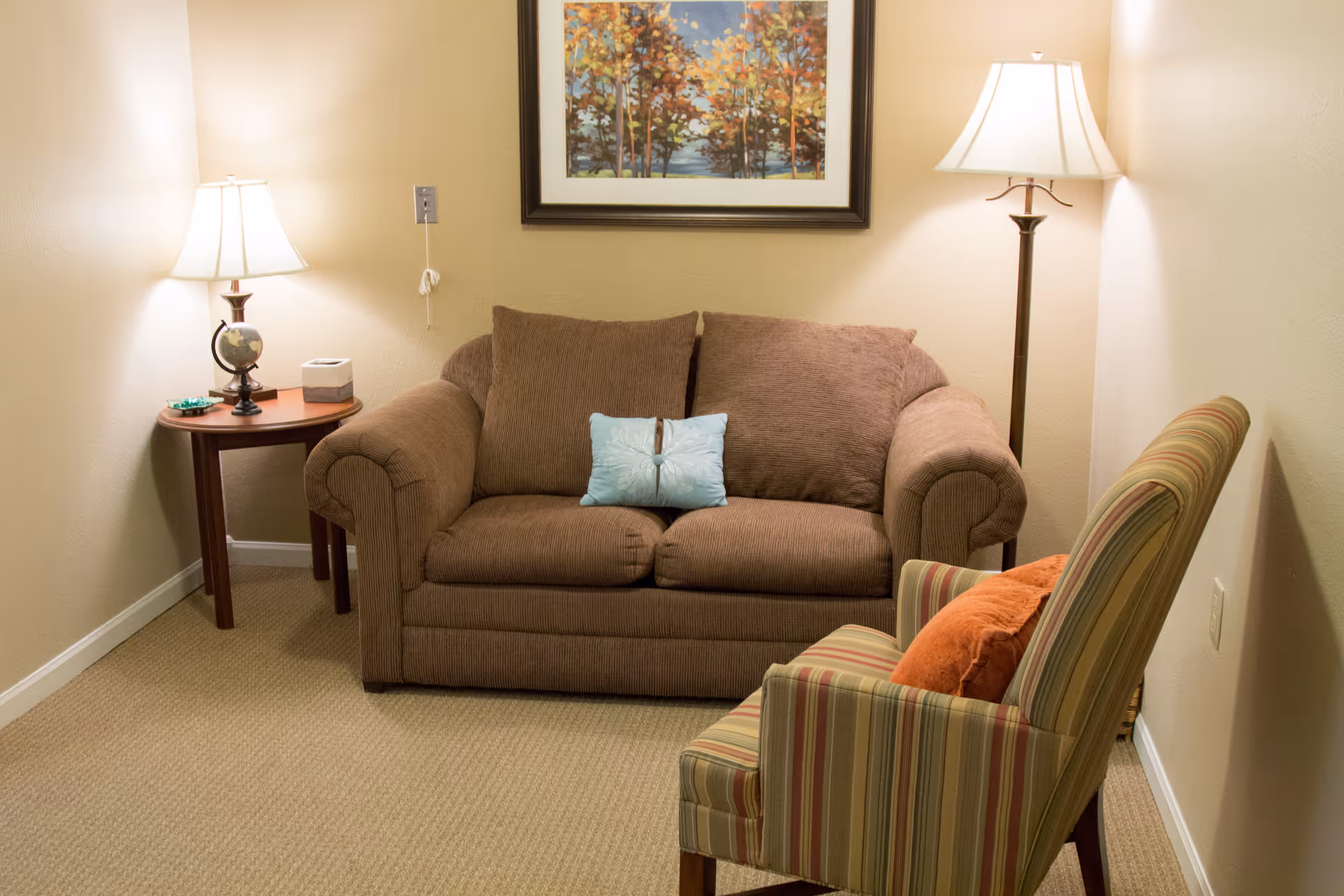 Small living room with a brown loveseat, a striped armchair, a side table with a lamp, a floor lamp, and framed artwork on the wall.