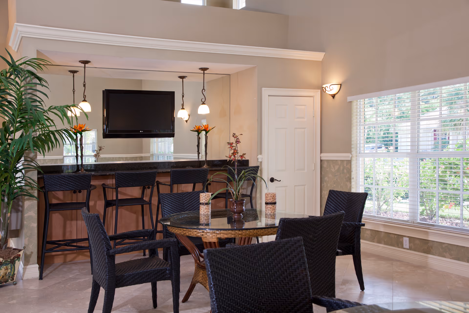 A bright and cozy common area with a round glass-top table surrounded by black wicker chairs. Behind the table is a counter with four high black wicker chairs, a mounted flat-screen TV, and three hanging pendant lights. There are decorative plants on the counter and a large window with white blinds letting in natural light. The walls are painted beige with white trim, and there is a white door in the corner.