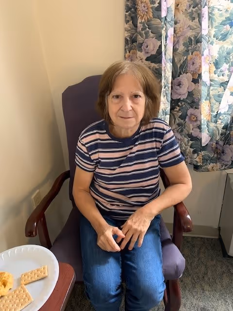 An elderly woman with short brown hair wearing a striped t-shirt and blue jeans is sitting in a purple armchair next to a small table with a plate of crackers and cheese. Behind her is a floral curtain and a beige wall.