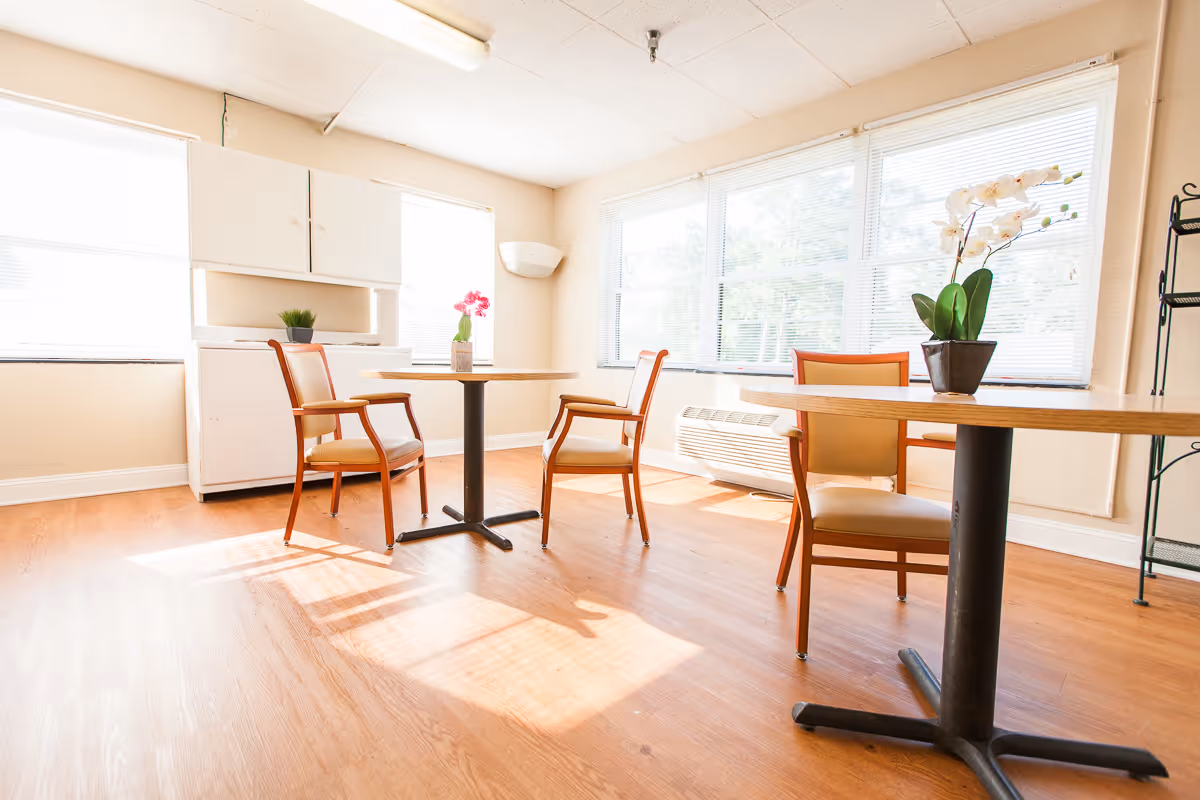 A bright and airy dining area with wooden floors, two round tables each with two chairs, and potted plants on the tables. Large windows with blinds allow natural light to fill the room. A white cabinet and a wall-mounted heater are visible in the background.