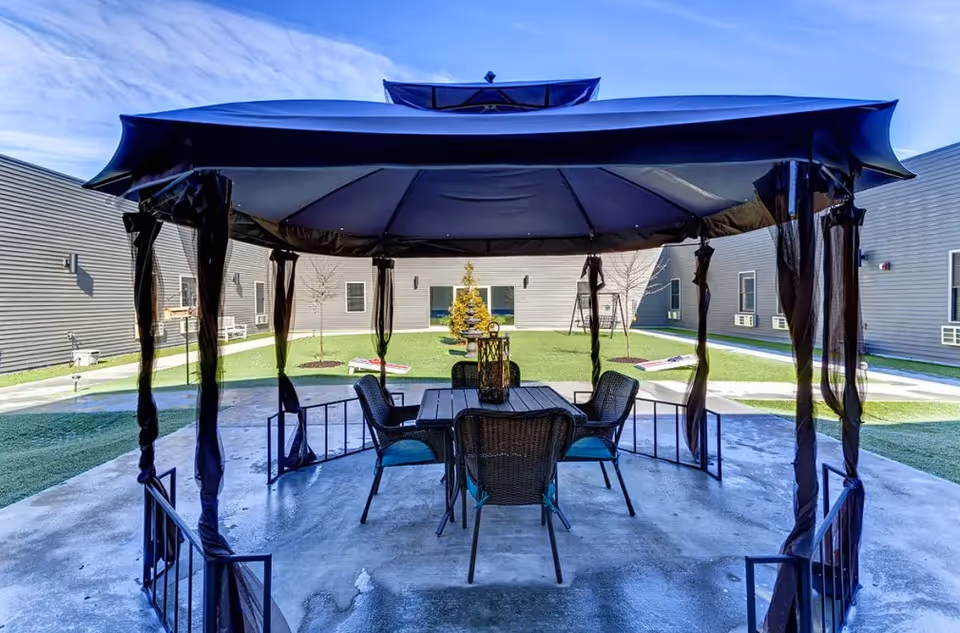 Covered gazebo with a table and chairs in the center of a grassy residential courtyard surrounded by single-story building wings.