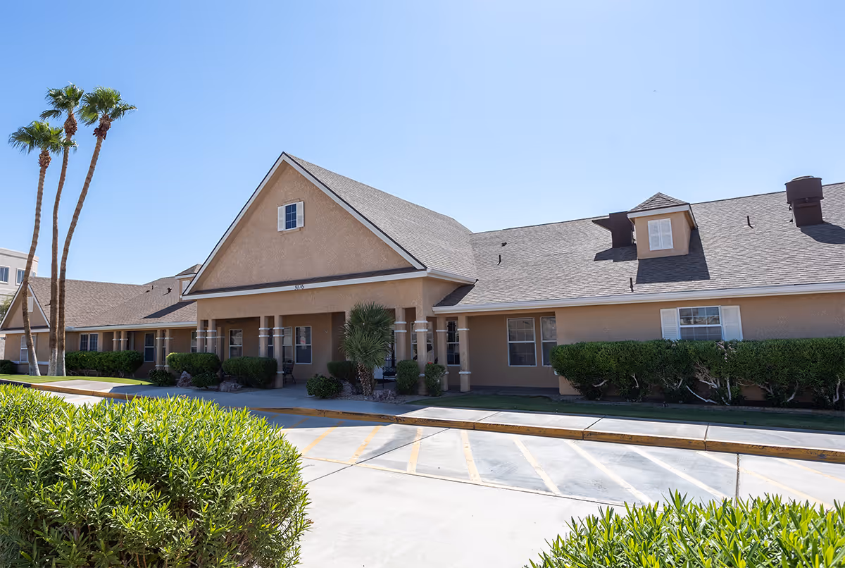 Exterior view of a single-story senior living facility building with a beige facade, multiple windows, and a peaked roof. There are palm trees and green bushes in front of the building under a clear blue sky.