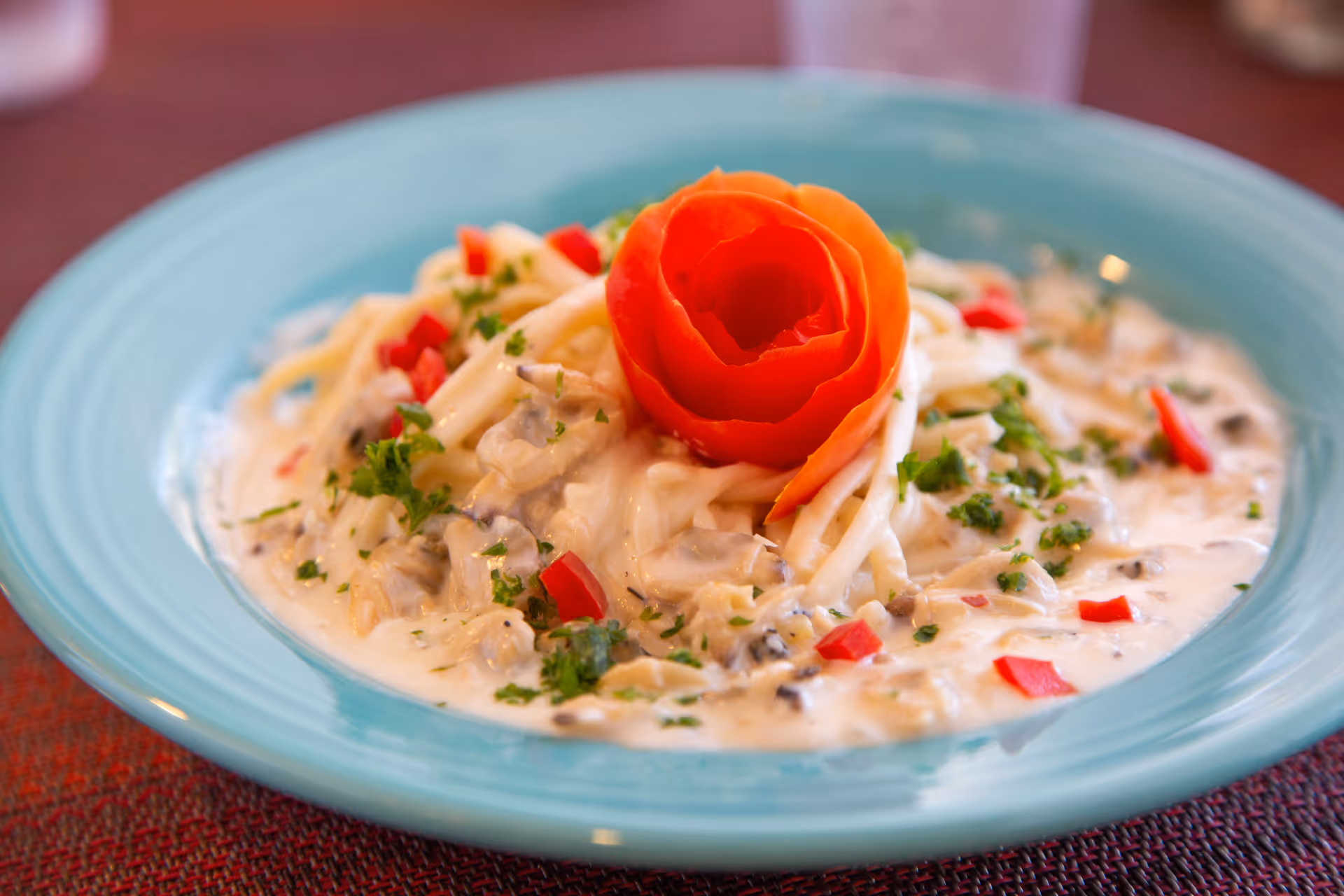 A plate of creamy pasta garnished with chopped herbs and red bell pepper pieces, topped with a decorative tomato rose, served on a light blue plate.