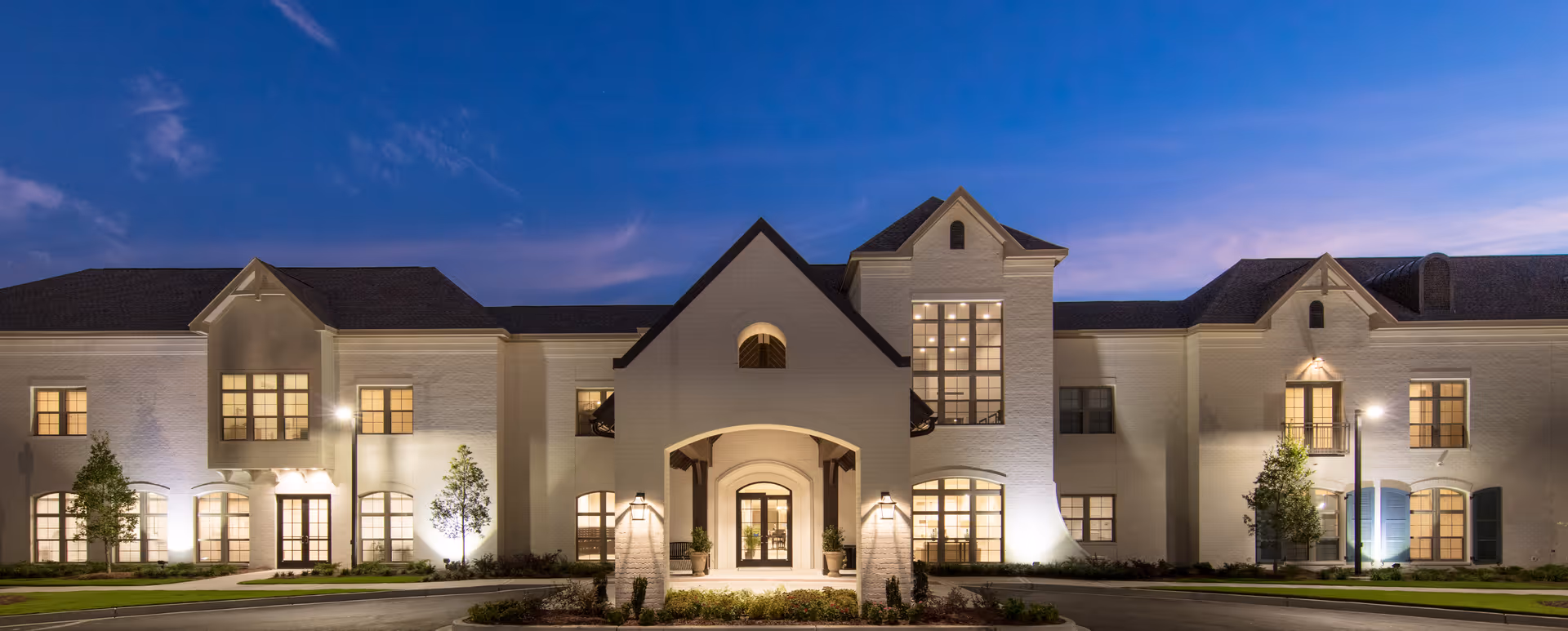Front exterior of a large white senior living building illuminated at dusk with a central arched entrance.