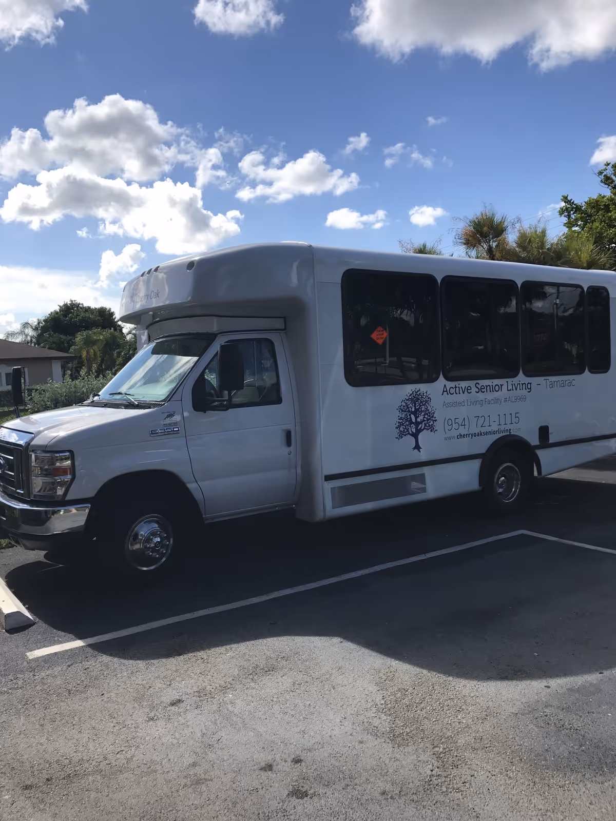A white shuttle bus parked in a parking lot under a partly cloudy blue sky. The bus has signage on the side that reads 'Active Senior Living - Tamarac' with contact information and a tree logo.