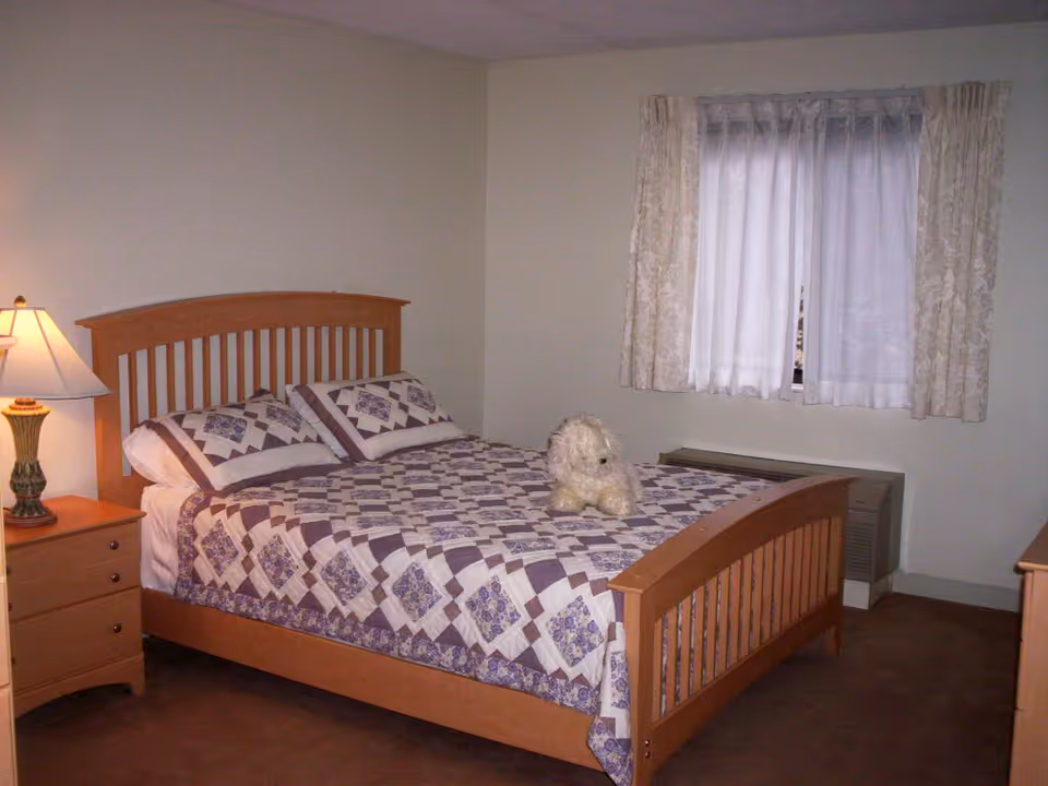A simple bedroom with a wooden bed topped with a patterned quilt and a small stuffed dog, a nightstand with a lamp, and a curtained window.