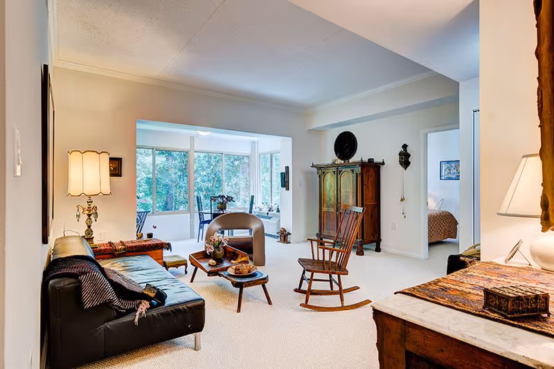 A cozy living room with a black leather sofa, a wooden rocking chair, a small coffee table with decorative items, and a vintage wooden cabinet. The room is well-lit with natural light coming through large windows in the adjacent sunroom area. A bedroom is visible through an open doorway.