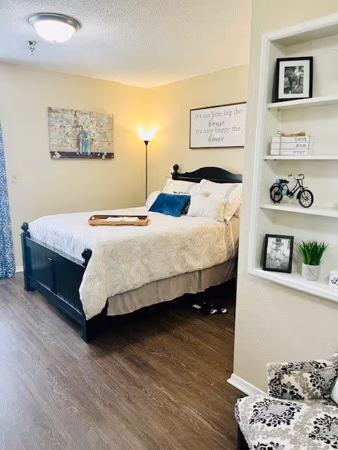 A cozy bedroom with a black wooden bed frame and cream-colored bedding. There is a wooden tray on the bed, a floor lamp in the corner, and wall art above the bed. To the right, there is a built-in shelf with decorative items including framed photos, books, a small bicycle model, and a potted plant. A patterned chair is partially visible in the foreground.