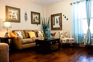 A cozy living room featuring a comfortable sofa with decorative pillows, a coffee table, and framed artwork on the walls. Natural light streams in through a window with light blue curtains.