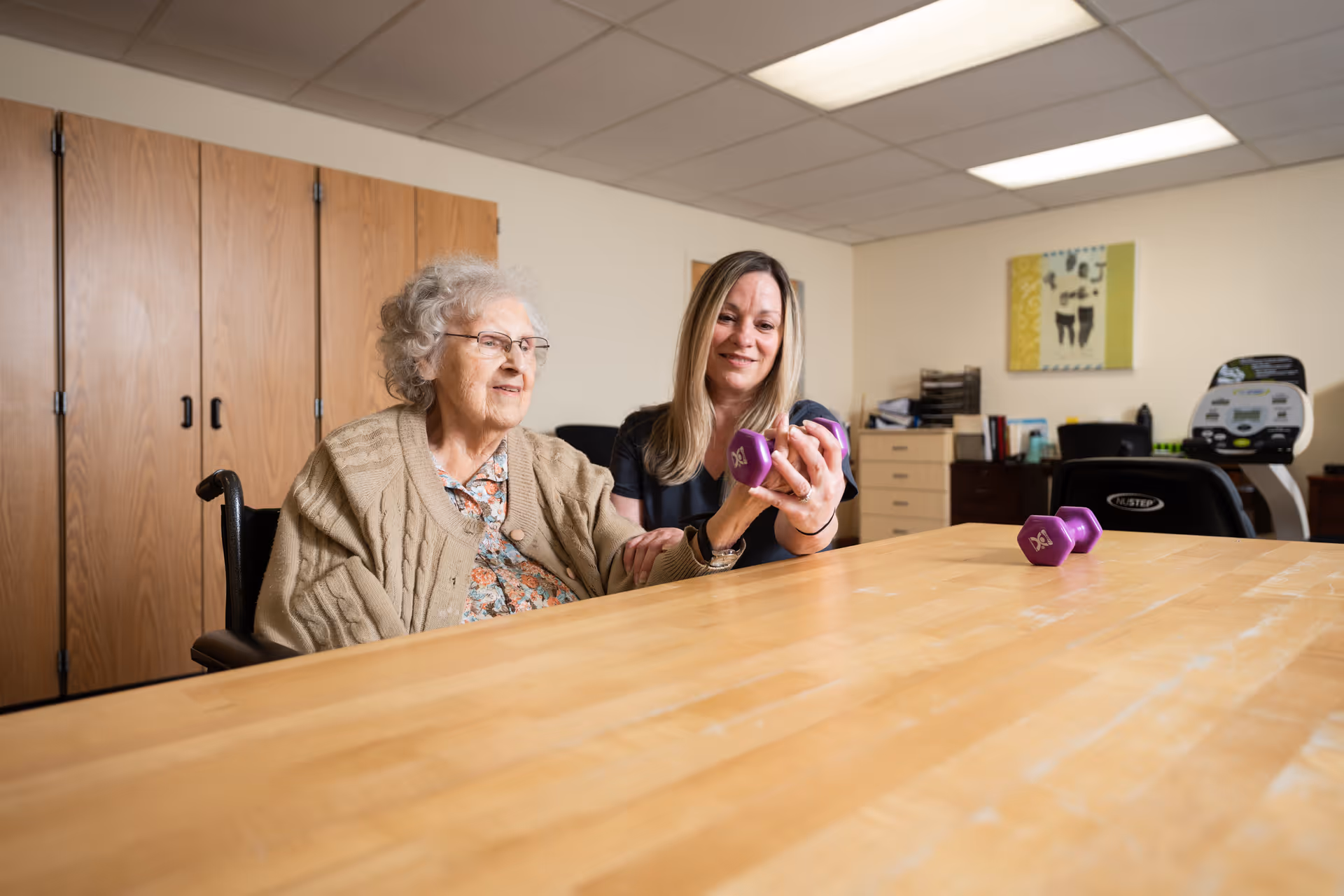 An elderly woman sitting in a wheelchair at a wooden table is assisted by a younger woman who is helping her lift a purple dumbbell. The room has wooden cabinets, a treadmill, and office furniture in the background.