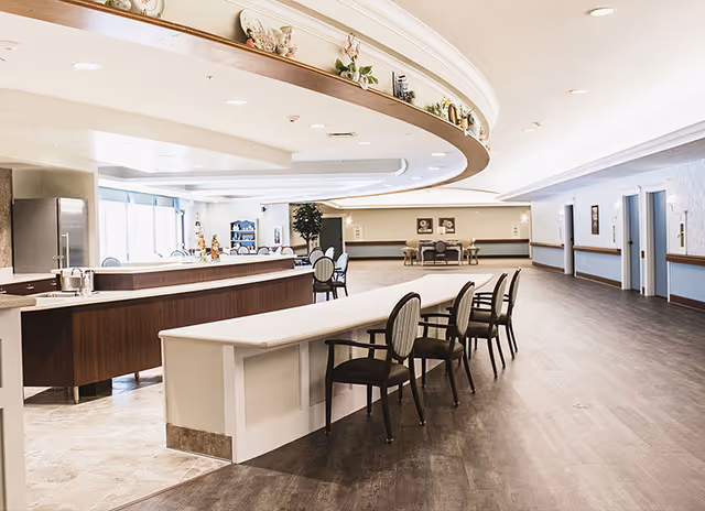 A spacious, well-lit interior common area in a senior living facility featuring a long counter with several chairs, a kitchen area with a refrigerator and sink, and additional seating and tables in the background. The ceiling has a curved design with decorative plants and items on a ledge above the counter.