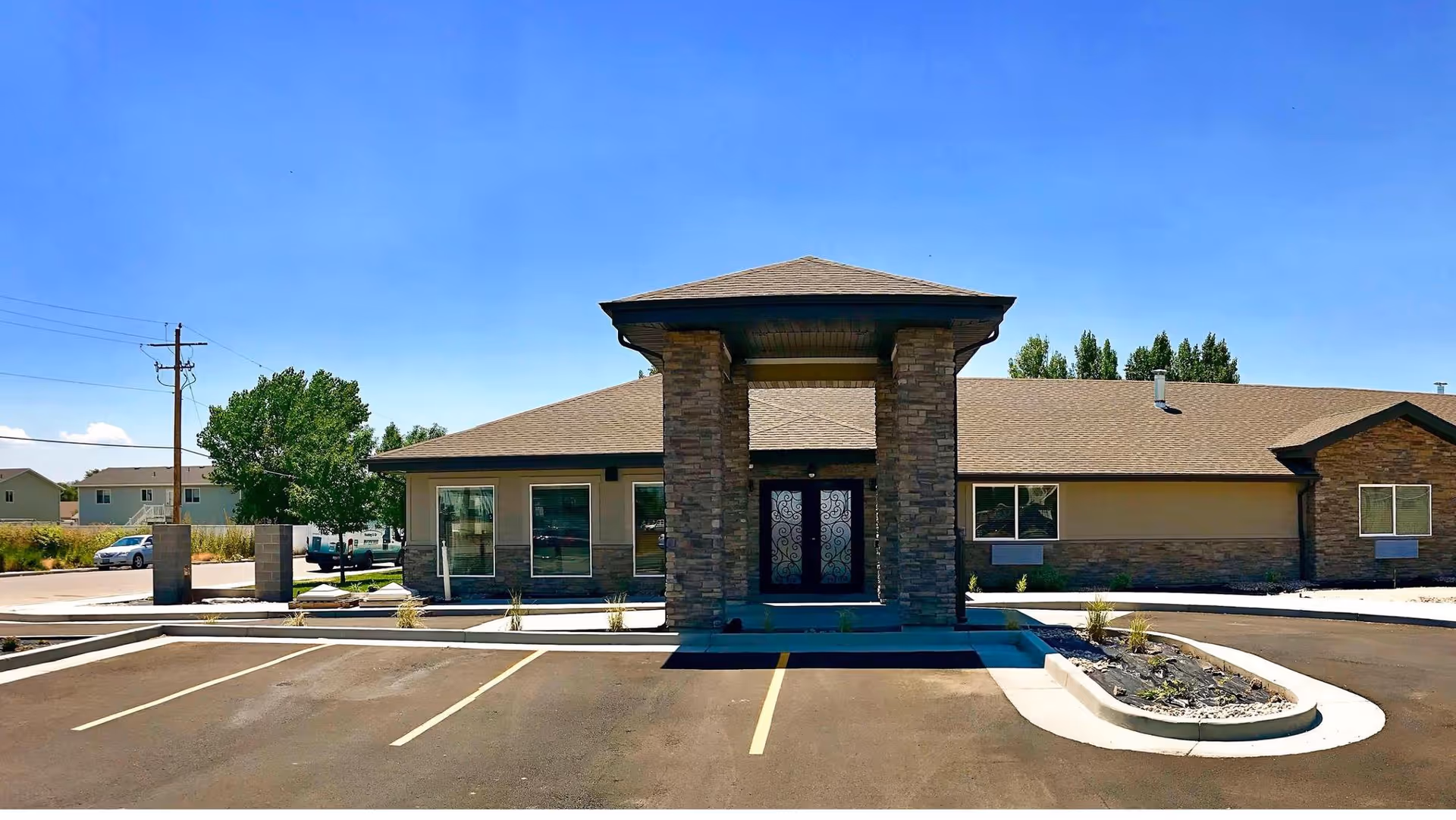 Exterior front view of a single-story assisted living facility building with a covered entrance supported by stone pillars, parking spaces in front, and clear blue sky above.