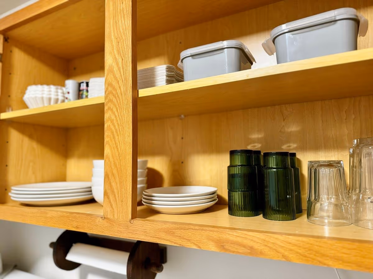 Wooden kitchen shelves holding white plates, bowls, green and clear drinking glasses, gray storage containers, and some small cups and dishes.