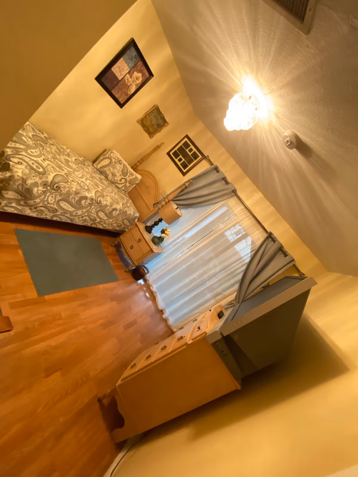 A cozy bedroom with a single bed covered in a patterned bedspread, a wooden nightstand with a lamp and decorative items, a dresser with a television on top, light blue curtains over a window, wooden flooring, and framed artwork on the walls.