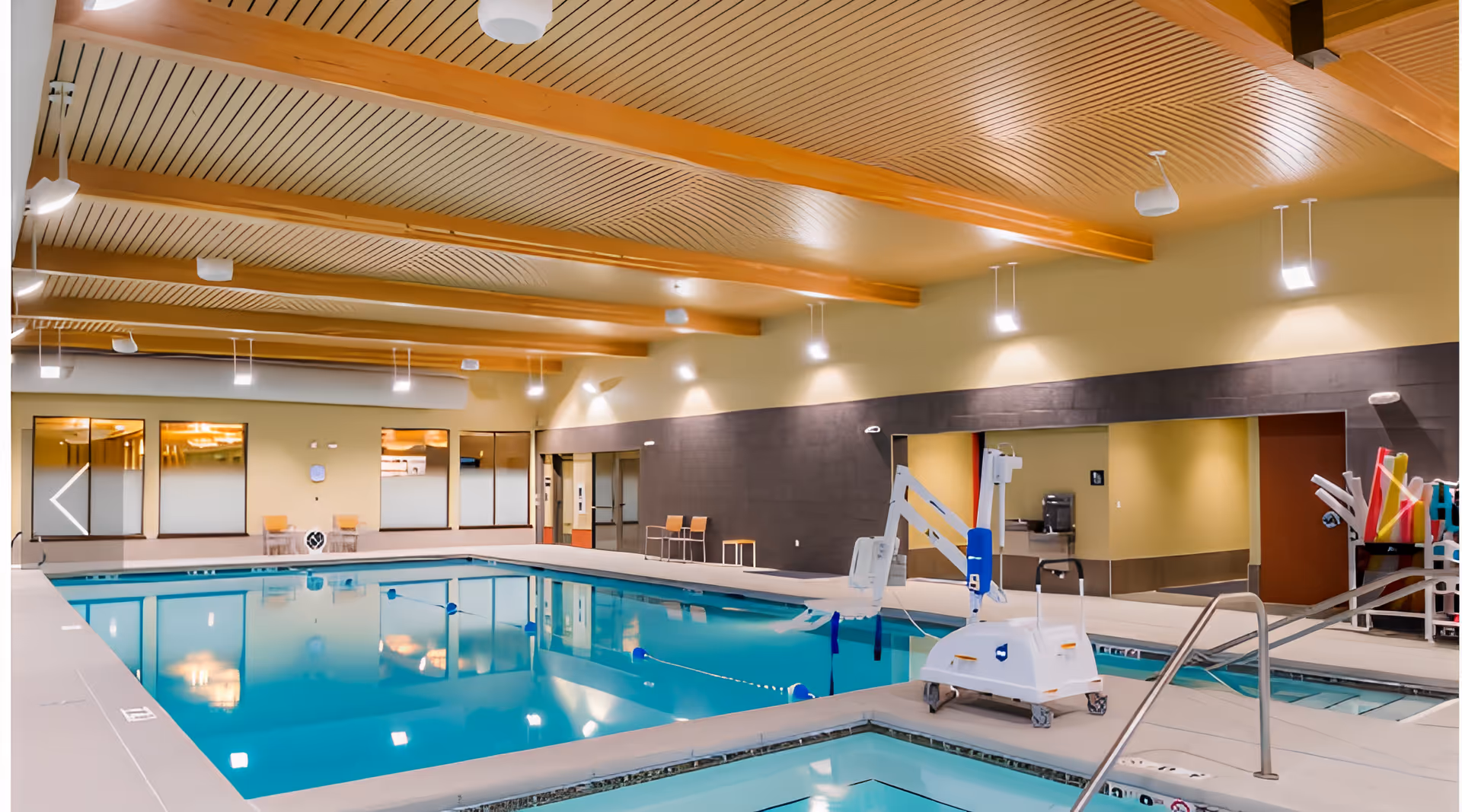Indoor swimming pool area with a smaller adjacent pool, equipped with a pool lift for accessibility. The ceiling has wooden beams and modern lighting fixtures. There are chairs and tables along the walls, and pool safety equipment is visible.