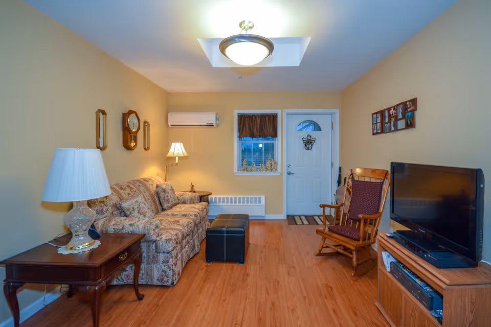 A cozy living room with a floral patterned sofa, a wooden rocking chair with a cushion, a wooden side table with a lamp, a wall clock, and a flat-screen TV on a wooden TV stand. The room has light yellow walls, a wooden floor, a window with a brown valance, and a white front door with a decorative hanging.