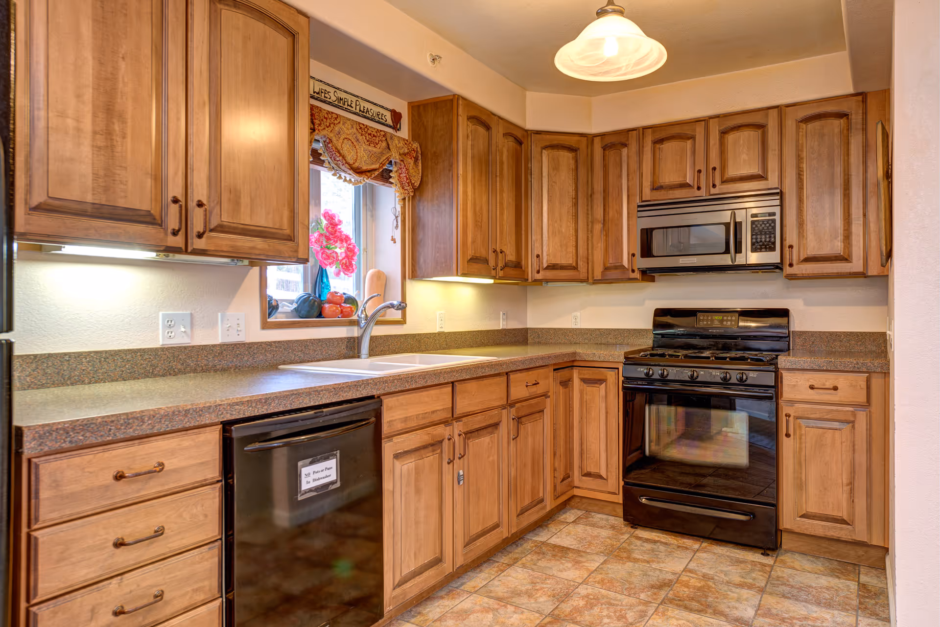 A kitchen with wooden cabinets, a black stove with an oven, a microwave above the stove, a black dishwasher, a double sink under a window with a floral valance, and a countertop with various fruits and a vase with pink flowers.