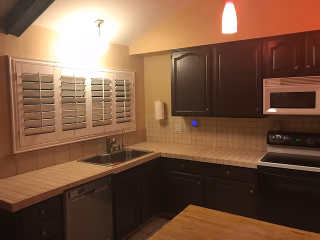 Interior view of a kitchen with dark wooden cabinets, a tiled countertop, a stainless steel sink under a window with white shutters, a dishwasher, an electric stove with an oven, and a microwave above it. The lighting includes a ceiling light and a red pendant light.