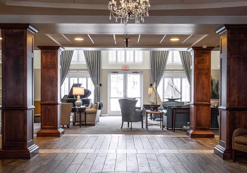 Spacious and elegant living room area with wooden columns, a chandelier, comfortable seating including armchairs and sofas, a grand piano, decorative model sailboats, large windows with curtains, and a glass door leading outside.