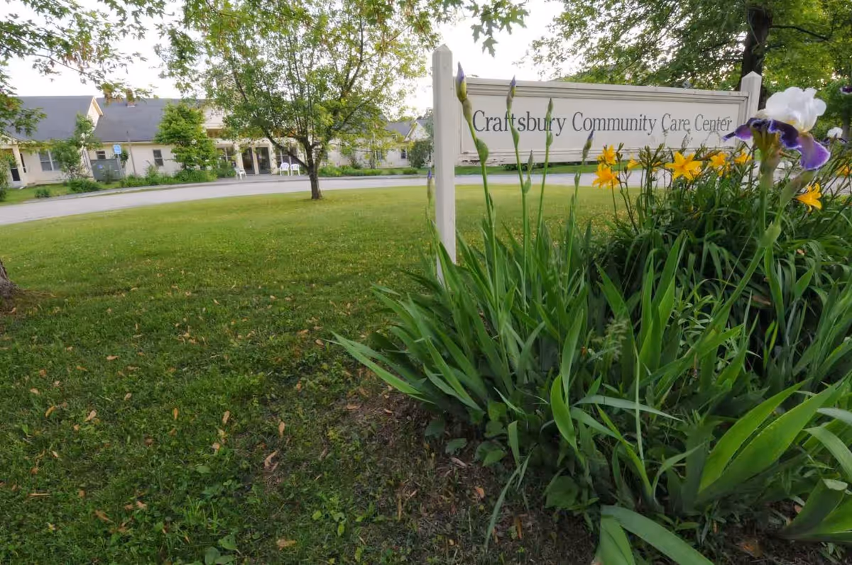 Outdoor view of the Craftsbury Community Care Center sign surrounded by green plants and flowers, with the facility building and trees visible in the background.