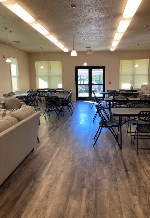 Interior view of a common area in Elmwood Manor with multiple tables and black folding chairs arranged neatly. There are pendant lights hanging from the ceiling and large windows with blinds letting in natural light. A beige couch is visible on the left side, and a glass door at the far end leads outside.