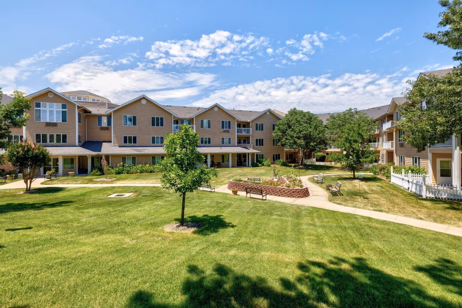 Exterior courtyard view of a multi-story senior living building with a grassy lawn, trees, walkways, and benches.