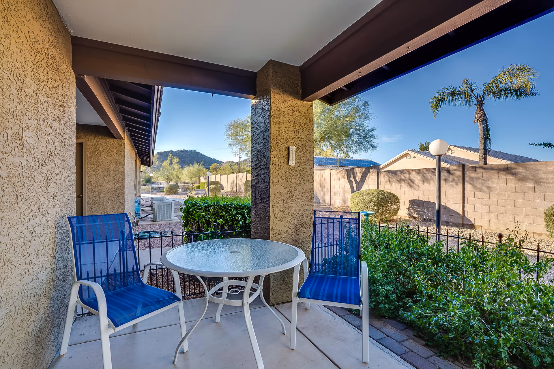 Covered patio with a round glass table and two blue chairs overlooking a landscaped yard with palm trees and distant hills.