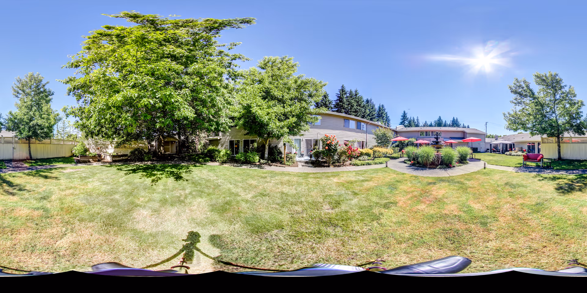 Sunny landscaped courtyard with lawn, trees, shrubs, and patio seating with red umbrellas in front of a two-story building.