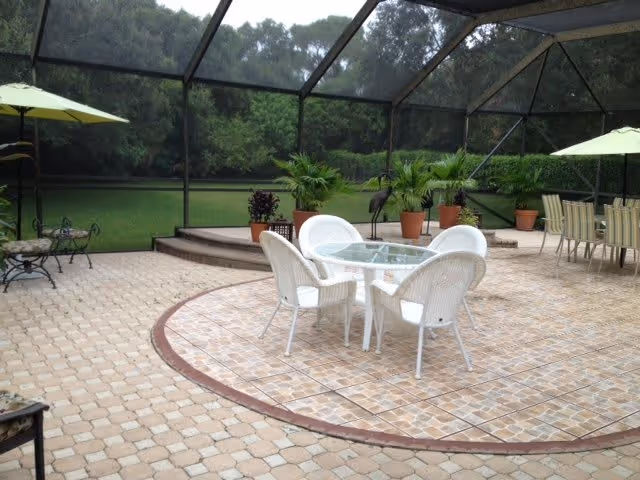 A screened-in outdoor patio area with a round glass table surrounded by four white wicker chairs. There are several potted plants and a metal bird sculpture on a raised platform in the background. Additional seating with a table and chairs is visible to the right, and a green umbrella shades a bench on the left side. The patio is paved with light-colored tiles and bordered by a red circular trim.