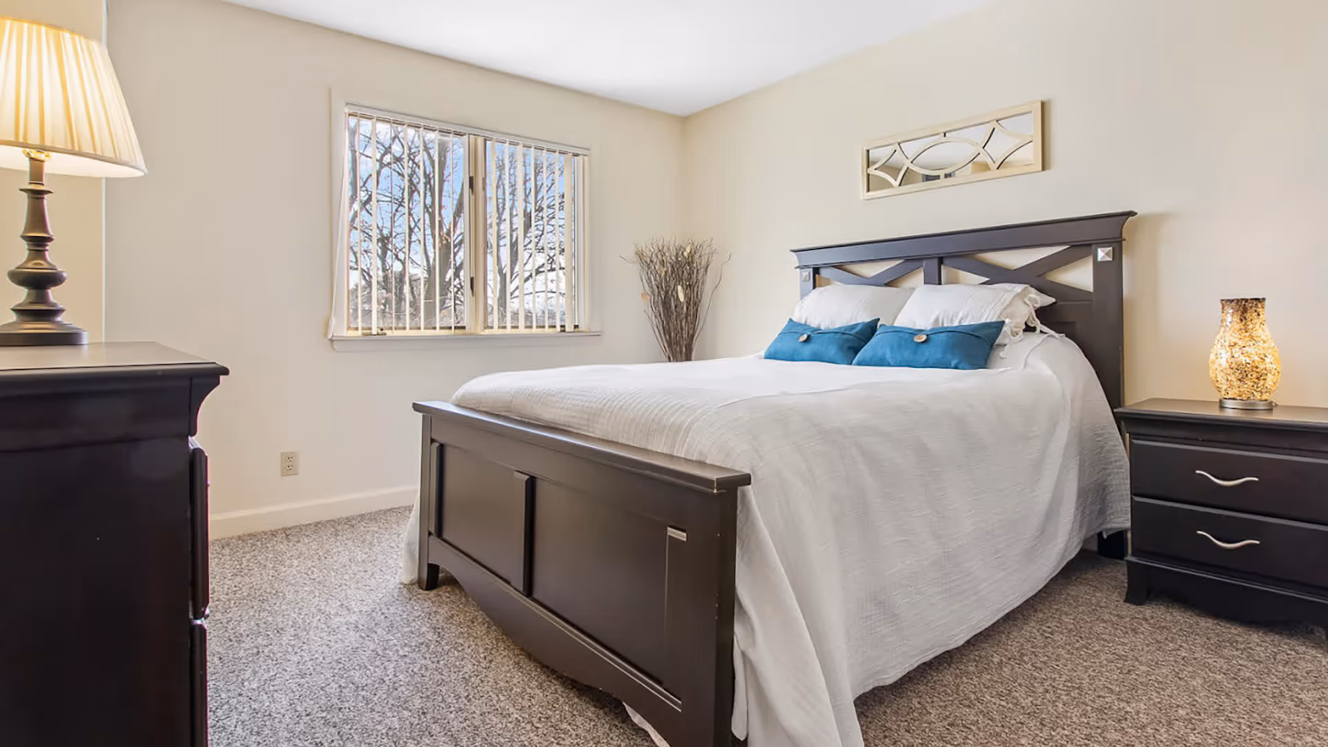 A bright bedroom with a large bed featuring a dark wooden frame, white bedding, and two blue accent pillows. There is a window with vertical blinds letting in natural light, a nightstand with a decorative lamp, a dresser with a lamp, and a decorative mirror on the wall above the bed.
