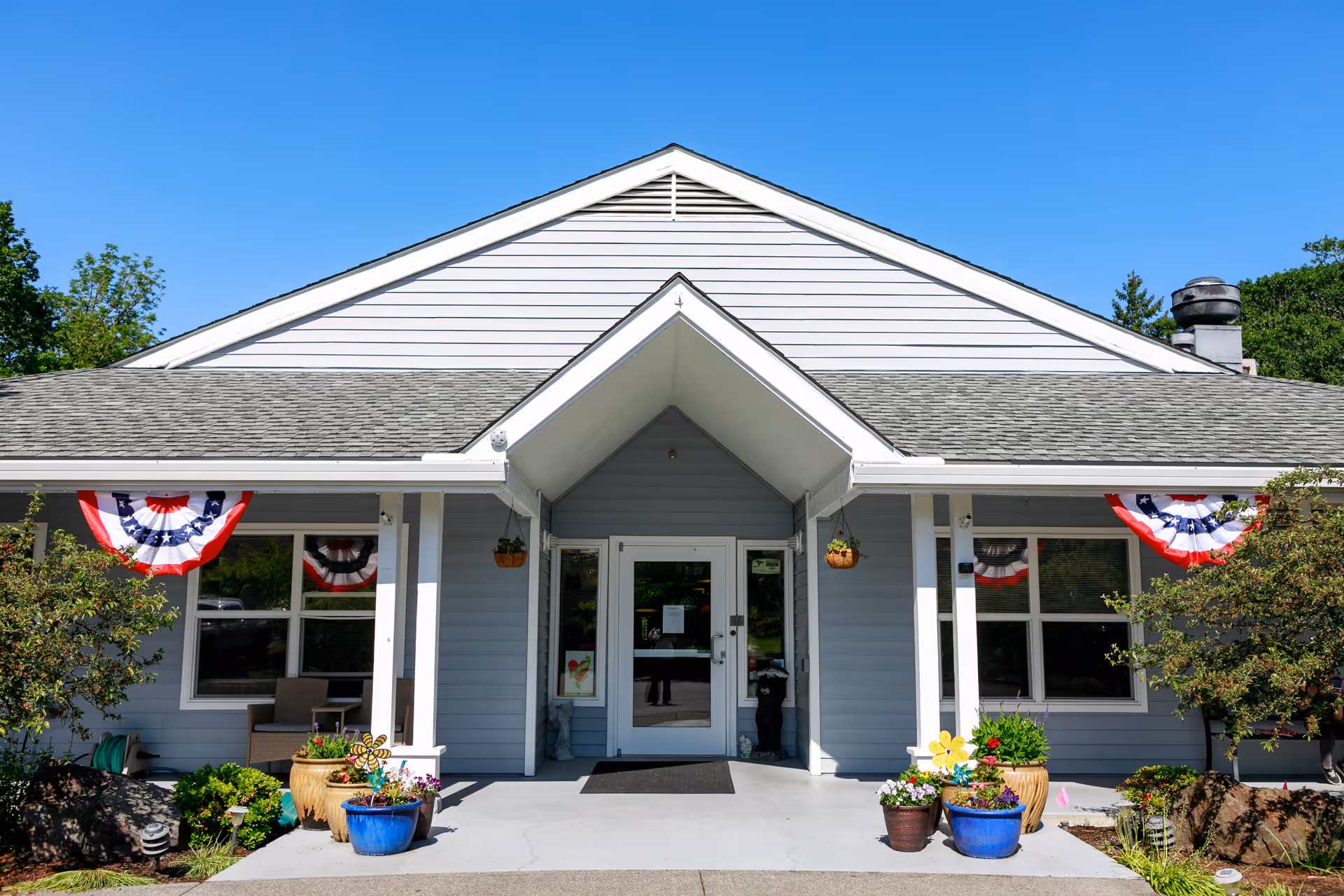 Front exterior view of a single-story building with light blue siding and a gray shingled roof under a clear blue sky. The entrance has a covered porch with two white pillars and glass double doors. There are several potted plants and flowers arranged symmetrically on either side of the entrance. Red, white, and blue patriotic bunting decorations hang above the windows on both sides of the porch.