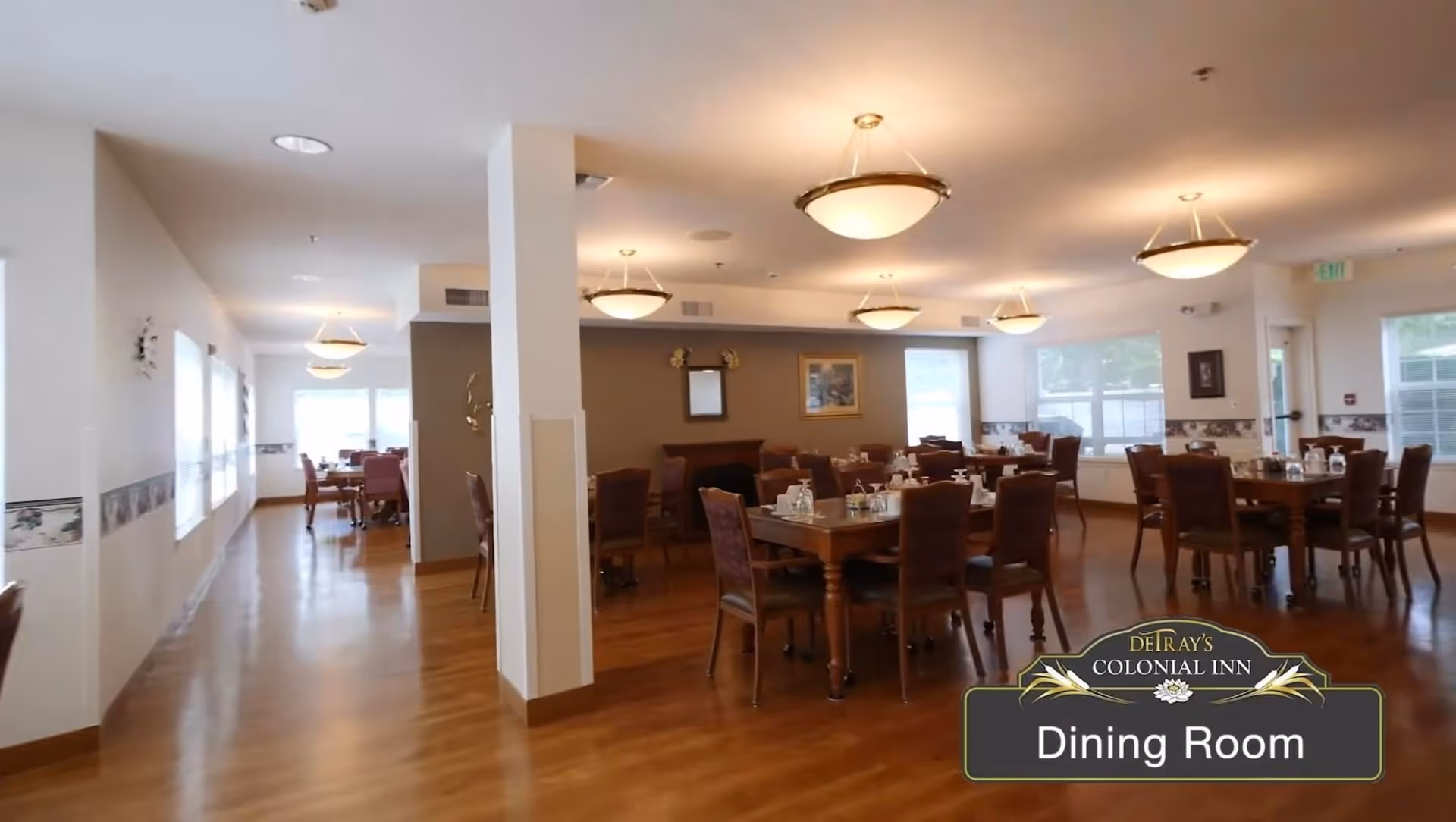 Spacious dining room with multiple wooden tables and chairs arranged neatly. The room has warm lighting from ceiling fixtures and large windows letting in natural light. The floor is wooden, and the walls are decorated with framed pictures and a mirror. A sign in the bottom right corner reads 'DeTray's Colonial Inn Dining Room'.