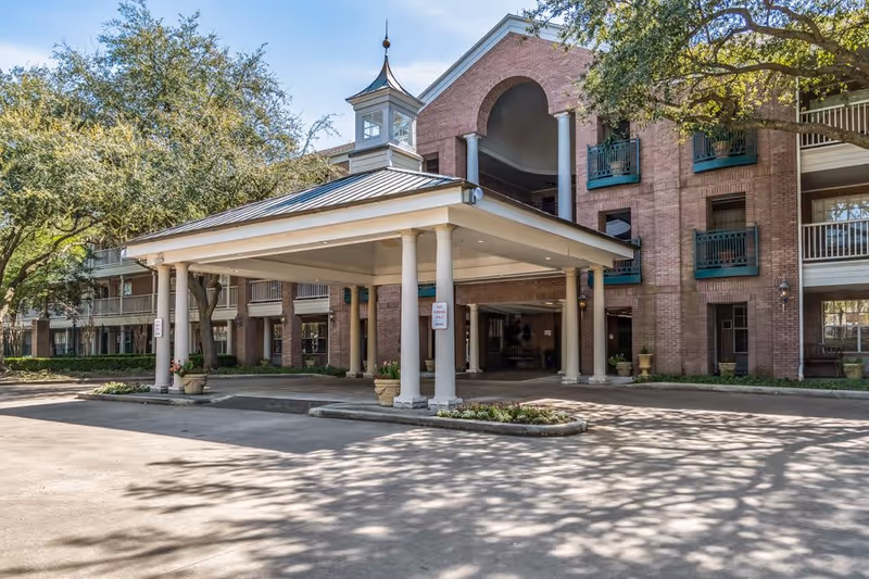 Exterior view of Elison Park Independent Living facility showing a covered entrance with columns, a brick building with balconies, and surrounding trees casting shadows on the driveway.
