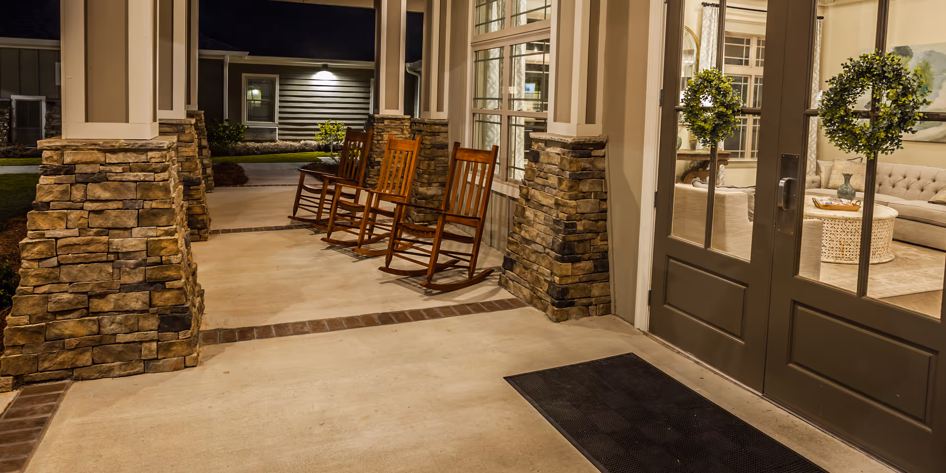Covered outdoor patio area at night with stone pillars and three wooden rocking chairs lined up along the wall. Through glass doors, a cozy indoor seating area with a beige tufted sofa, round woven coffee table, and two potted topiary plants is visible.
