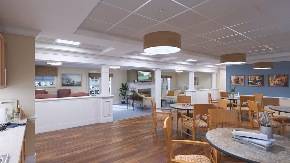 A spacious senior living common area with wooden floors and round tables surrounded by wooden chairs. The room features a cozy sitting area with sofas and armchairs near a fireplace and a wall-mounted TV. The walls are decorated with paintings and the ceiling has modern circular light fixtures.