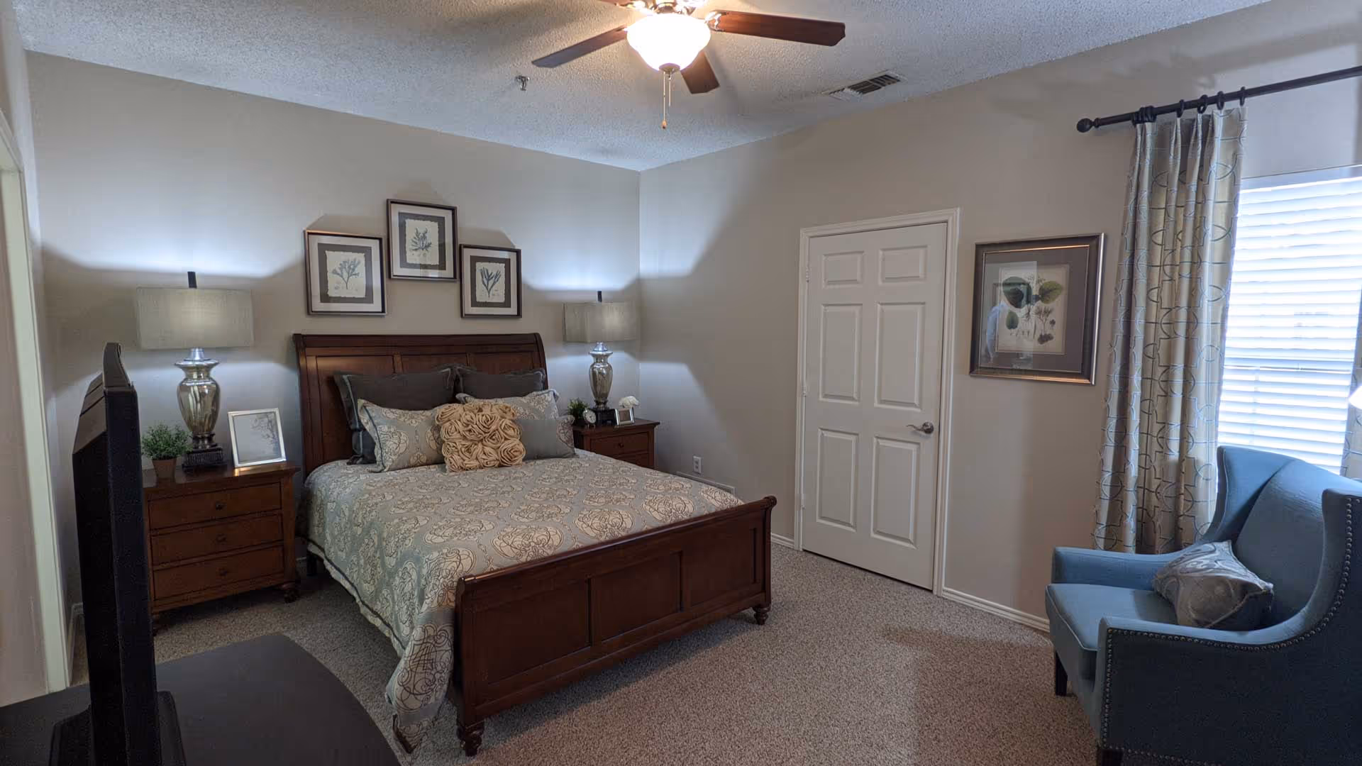 A cozy bedroom with a wooden bed frame and a patterned bedspread. There are multiple pillows on the bed, two bedside tables with lamps, framed botanical artwork above the bed, a blue armchair with a decorative pillow near a window with patterned curtains, and a ceiling fan with a light fixture.