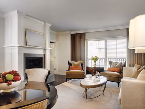 A bright living room with a fireplace, sofa, two armchairs, a coffee table, and a dining table with a bowl of fruit.